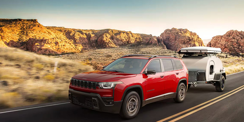 Red SUV towing a compact camper trailer along a scenic desert highway with rocky mountains in the background.